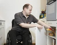 Ein Mann sitzt im Rollstuhl in der Küche. Er nimmt Essen aus einem Schrank.