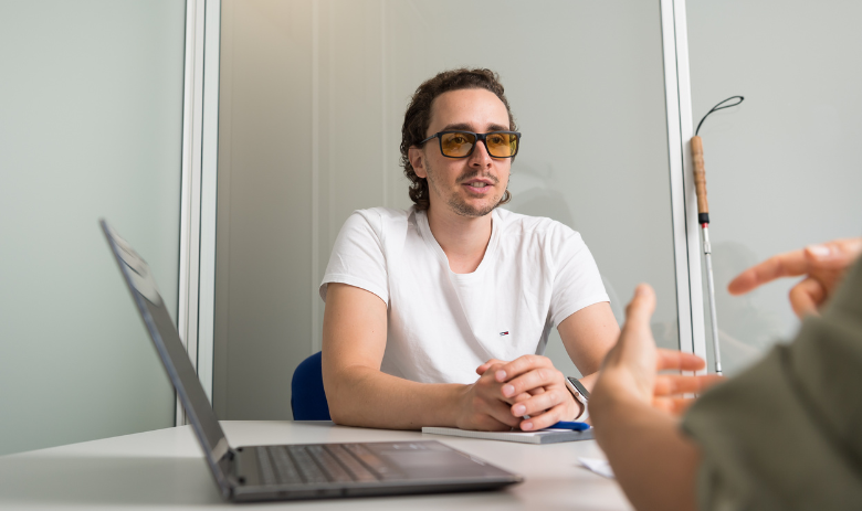 Barrierefreie Kommunikation Ein junger Mann mit Sonnenbrille hört einer Person ihm gegenüber zu. Neben ihm stehen ein PC und ein Blindenstock.